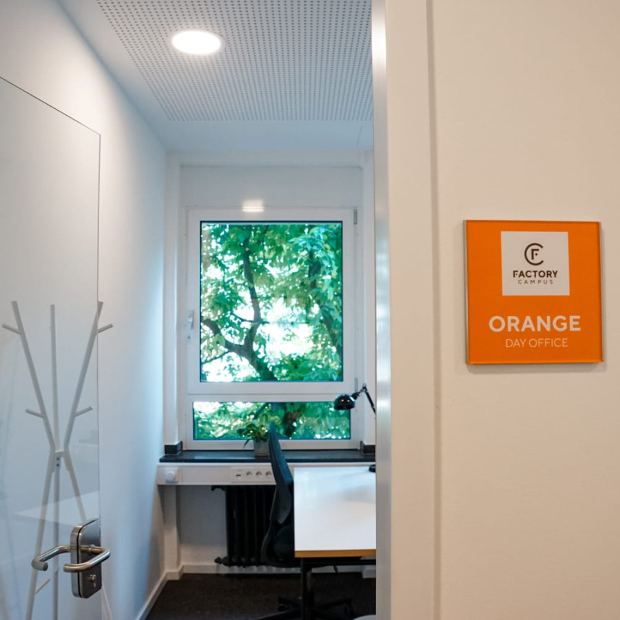 B&uuml;ro mit einem Schreibtisch, Kleiderst&auml;nder, orangfarbenem Teppich, Schreibtischstuhl, Tischpflanze und Bildschirm