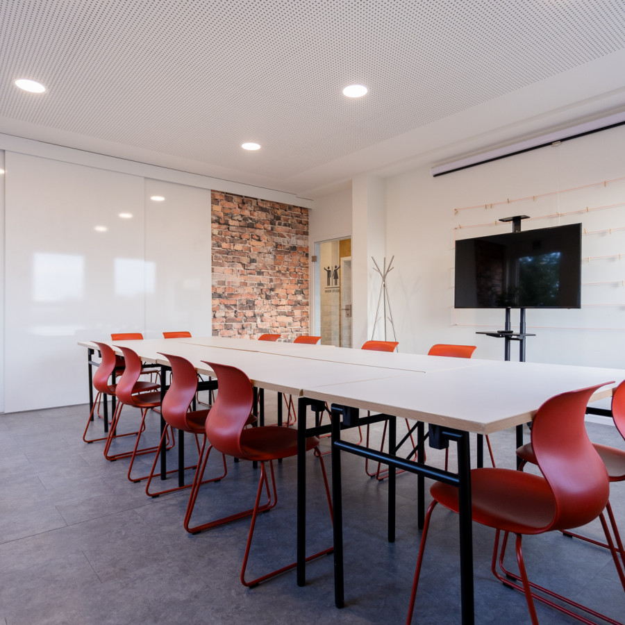 Konferenzraum "Selfie" mit langer wei&szlig;er Tafel, Pr&auml;sentationsbildschirm und roten St&uuml;hlen